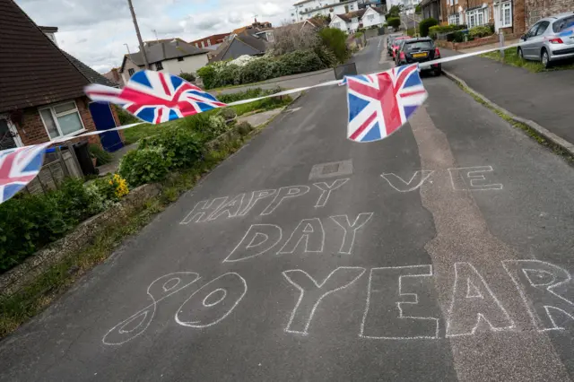 Union Jack flags are seen above a chalk written message on the road which says "Happy VE Day 80 years'
