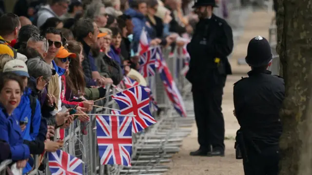 Crowds stand behind metal barriers waving UK flags as police officers look on from the other side