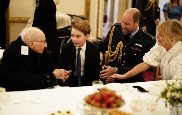 Prince George speaks to elderly man at table with Prince William behind him