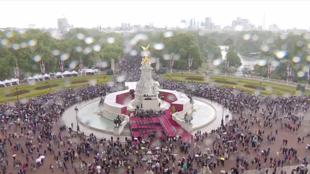 Rain drops appear on camera with crowds below, outside Buckingham Palace, holding umbrellas