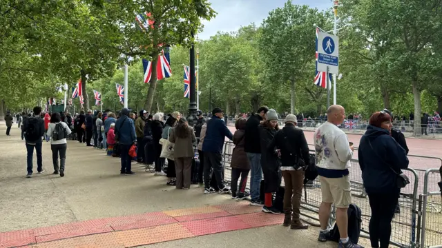 Line of people standing along The Mall behind metal barriers