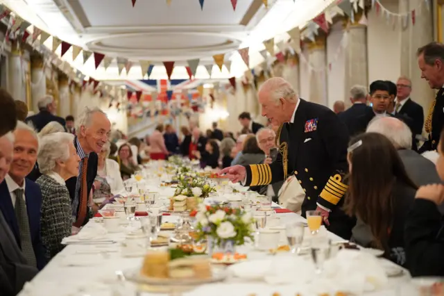 King Charles leans over dinner table and points his hand towards people on the other side