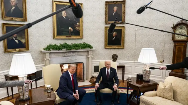 Trump and Biden are seated together in the Oval Office. There is no gold on the fire place, but there are pictures on the wall