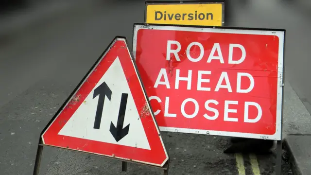 A road ahead closed sign which is square with white letters on a red background. There is a red triangle sign with two black arrows to show a two way street and behind you can see the top of a yellow diversion sign.