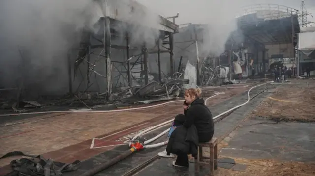 A vendor sits outside the market as firefighters continue to extinguish the fire
