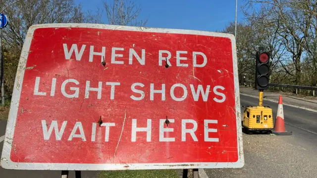 A red road sign which reads: "When red light shows wait here." Beyond it is a set of temporary traffic lights and a traffic cone.
