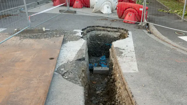 A hole in the road which has a blue plastic piping visible inside it. Surrounding the hole is metal fencing and low orange and white plastic bollards while the edge of a pavement is also seen at the top of the image.