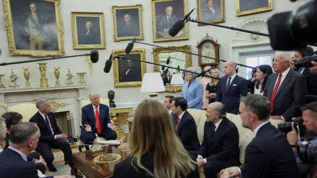 Trump and Carney sit in the Oval Office, while Trump officials and the media sit and stand around them, looking on. The fireplace is ornamented with gold, and there are pictures on the wall of former presidents