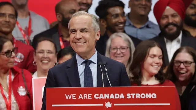 A smiling Mark Carney stands at a podium which has 'We are Canada Strong. Nous sommes un Canada fort' written on it. A group of smiling people behind him.