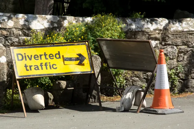 A diverted traffic sign in yellow with a black arrow on it pointing to the right. There is another sign seen from the back and a traffic cone on the pavement as well. In the background there is a low stone wall with yellow flowers growing out of it.