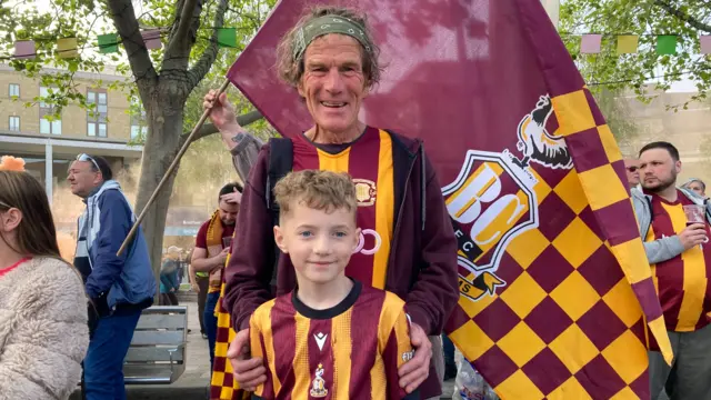 Man standing behind young boy both in Bradford tops with a Bradford flag behind them. They are both smiling.