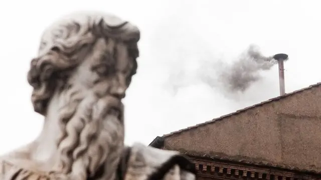 The head of statue is seen next to a chimeny emitting grey smoke