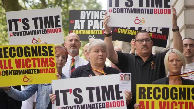 At least six campaigners can be seen holding up white, black and yellow background signs featuring slogans including ‘It’s time #contaminatedblood’