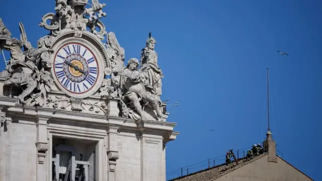 Firefighters work to fit a chimney on the roof of the Sistine Chapel. An ornate clock and a blue sky can be seen.