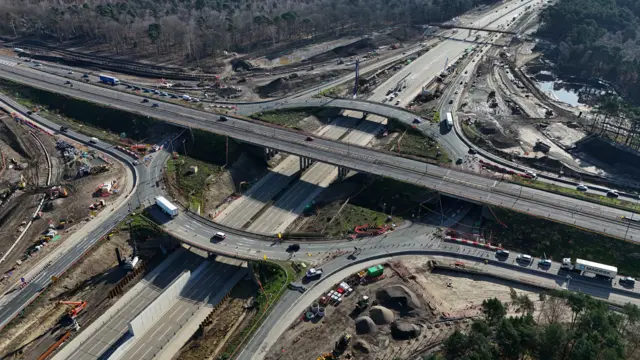 An aerial view of junction 10 where the M25 meets the A3. There are bridges over the road and cars queuing at different parts of the junction. Works are going on around the edge of the road with piles of earth seen and construction vehicles.