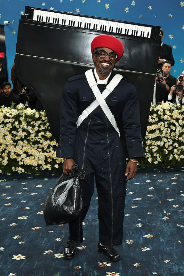 Andre 3000 wearing a black overall and a red hat. He is also wearing a black piano on his back.