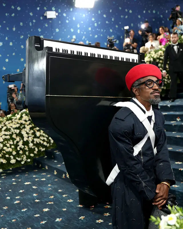 Andre 3000 wearing a black overall and a red hat. He is also wearing a black piano on his back.