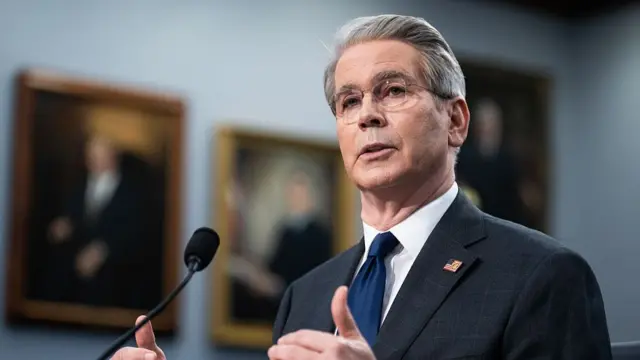 Scott Bessent, in a dark grey suit and blue tie, speaks in front of a microphone.