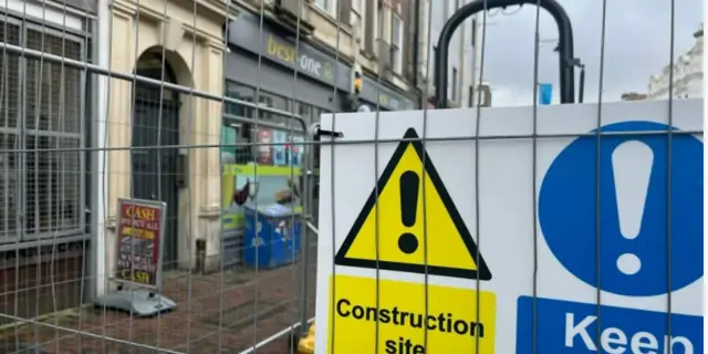 A metal fence with a sign attached which says "construction site" and has a yellow warning triangle on it. In the background there are shop fronts seen through the fencing.