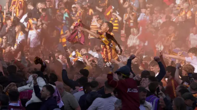 Footballer wearing claret and amber shirt hoisted aloft fans' shoulders celebrates