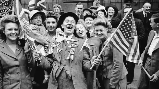 Black and white picture of people in London celebrating the end of the war. One man in a suit stands in the centre laughing while holding a US and a Union flag while a young woman leans on his left shoulder, other women surrounding them smiling
