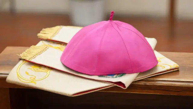 A pink skull cap and chasuble sit on a wooden bench