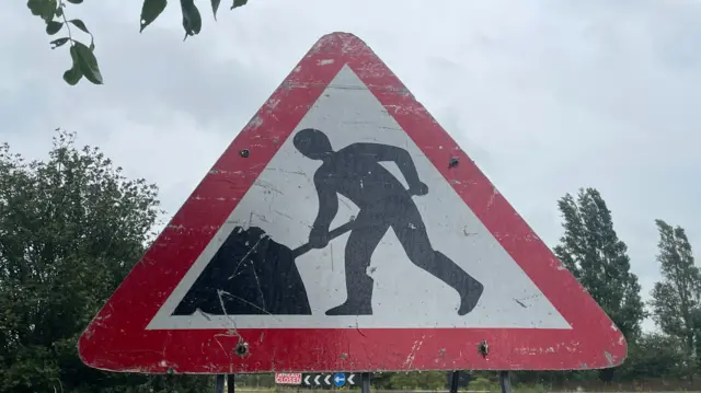 A roadworks sign which is a red triangle with a stick figure inside digging from a pile of earth. In the background there are trees and the sky visible.