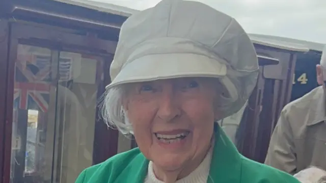 Dorothy Walsh, with grey hair, smiles at the camera. She is wearing a white baker boy hat and a green blazer.