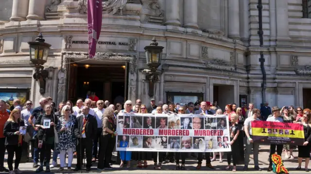 Victims and campaigners stand outside Central Hall in Westminster. Some hold banners and posters - a large one at the centre features images of people and reads ‘Murdered, Murdered, Murdered’.