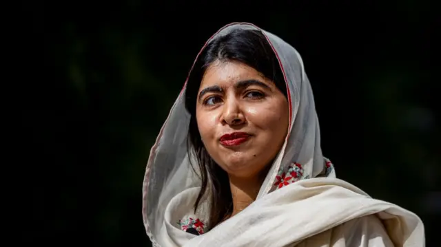 Malala pictured with a white headscarf loosely draped over her hear, with her hari and neck showing. the scarf has red trimming and red and green floral embroidery