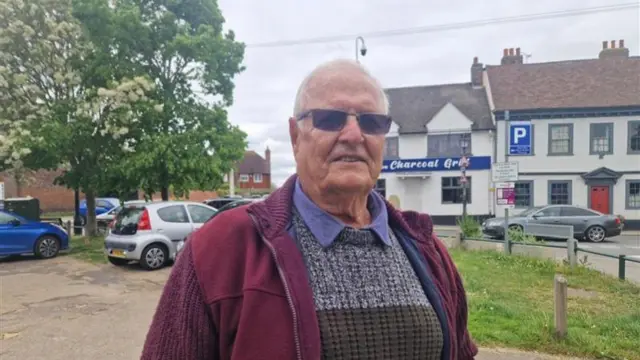 A man wearing a knitted jumper and sunglasses stood in a grassy car park. There are white buildings in the background.