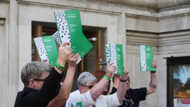 People hold up the green Infected Blood Inquiry report outside Central Hall in Westminster.