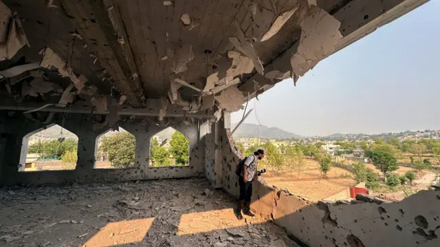 A man looks out a gaping hole in the wall of a damaged building. The ceiling is damaged. Outside, there are trees in the distance and clear skies.