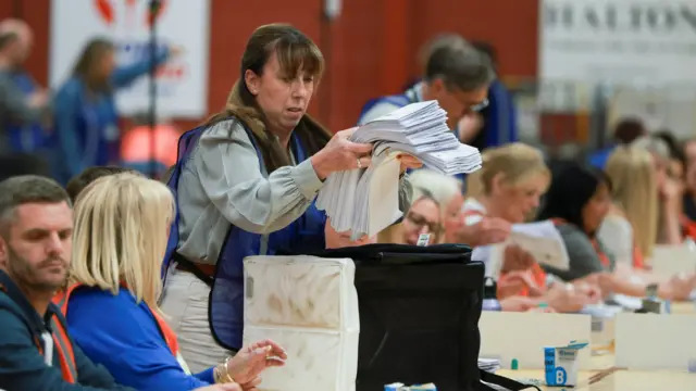 A woman holds a pile of ballots as people count ballots around her