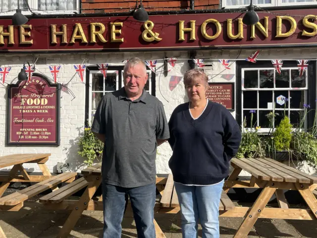 A man and a woman stand outside a white pub building. The pub has a red and gold sign reading The Hare & Hounds and picnic benches outside.