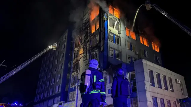 A building is seen on fire with emergency first responders looking on