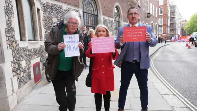 Campaigners outside the Infected Blood Inquiry at Church House, in Westminster, London, holding signs reading: 'Pay compensation now' and 'Time is not on our side. We are still dying. While they are still lying.'