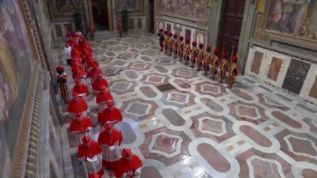 Cardinals walk together in a long line, each wearing red and white, through an elaborate chamber while guards in ceremonial dress stand by