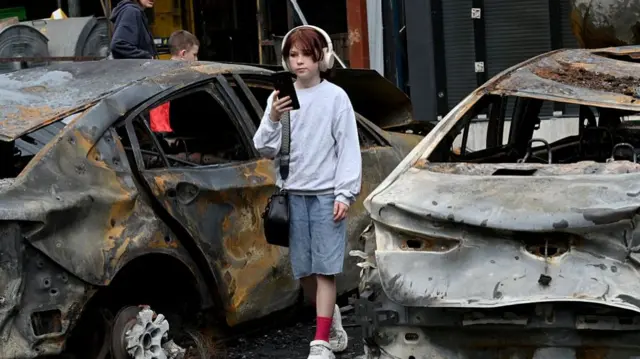 A girl walks between burnt cars