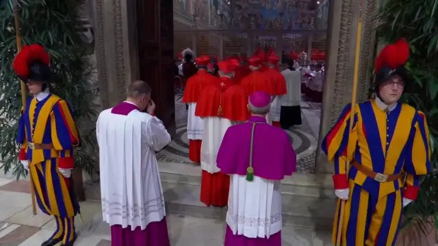 The cardinals, seen from the back, as the procession passes through a doorway, at which two guards stand side on either side in uniform