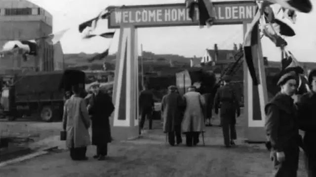 Flags fly from an archway with the words Welcome Home to Alderney across the top as soldiers and men dressed in coats stand nearby or walk towards the archway