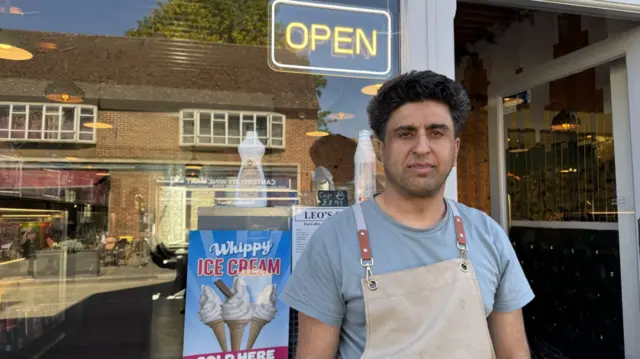 Ali Maroof, wearing a blue t-shirt and pale white dungarees, stands outside a cafe with an open sign and a picture of ice creams in the window