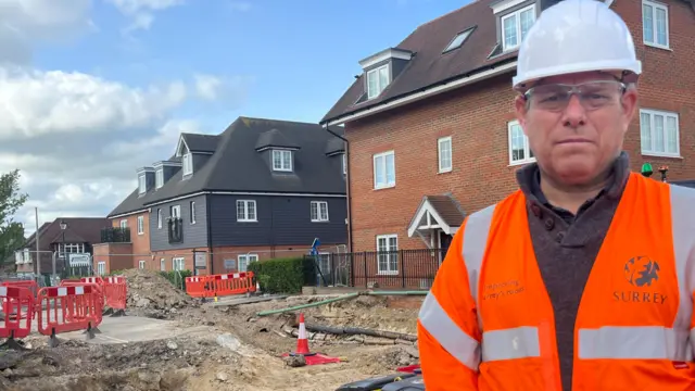 A man in an orange high-vis jacket and a white hard hat is stood beside an excavated hole. The hole is blocked with red construction barriers and is situated next to brick houses.