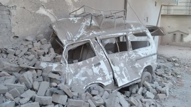 a destroyed vehicle against the wall of a building that is sitting on and surrounded by rubble. Everything is covered in grey dust/ash