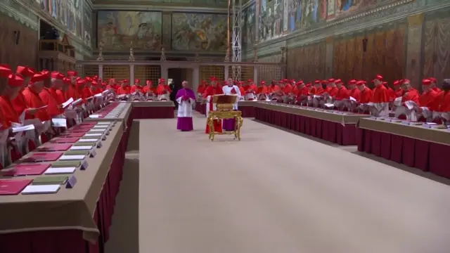 The cardinals, wearing red and white, stand at tables in the Sistine Chapel