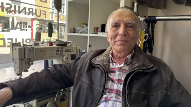 Sedat Ozdogan, wearing a red and white checked shirt and brown leather jacket, sits in his dry cleaning shop