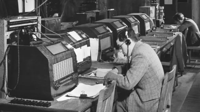 Black and white image of people wearing headsets sitting in front of radio speakers