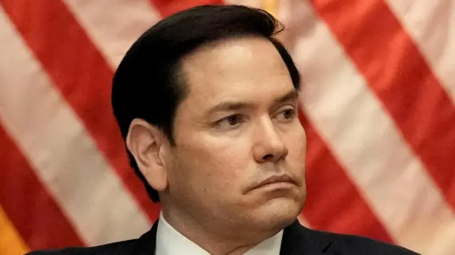 Marco Rubio looking to his left. He is wearing a Black suit over a white shirt.