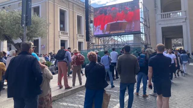 People gather around screens near St Peter's Basilica