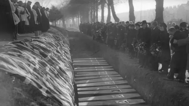 Black and white image of an open grave with a line of coffins